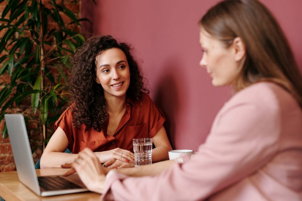 Two women having a friendly conversation in a cozy indoor setting with a laptop and coffee.